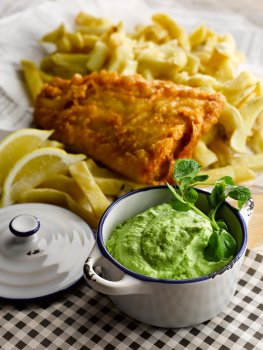 Food photograph close up of a cast iron pot filled with posh mushy peas garnished with watercress, in the background is a portion of golden battered cod and chips with lemon wedges served on newspaper